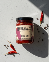 Jar of Fermentalists Chilli Jam on a concrete surface with red chilli peppers.
